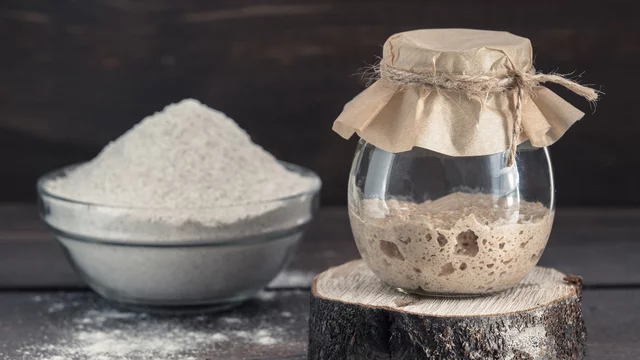 Auf dem Bild sieht man eine Glasschale mit Mehl und ein mit Papier verschlossenes Glas mit angesetztem Sauerteig.