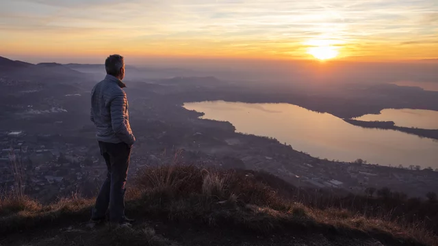 Ein Mann steht auf einer Anhöhe und schaut bei Sonnenuntergang ins Tal auf eine Ortschaft und zwei Seen. Abendstimmung.
