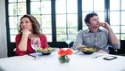 Ein Mann und eine Frau sitzen an einem Tisch mit Salat und Gläsern. Beide wirken nachdenklich und traurig, während sie getrennt voneinander in verschiedene Richtungen schauen. Ein kleines Blumenarrangement steht auf dem Tisch, das eine angenehme Atmosphäre schafft.