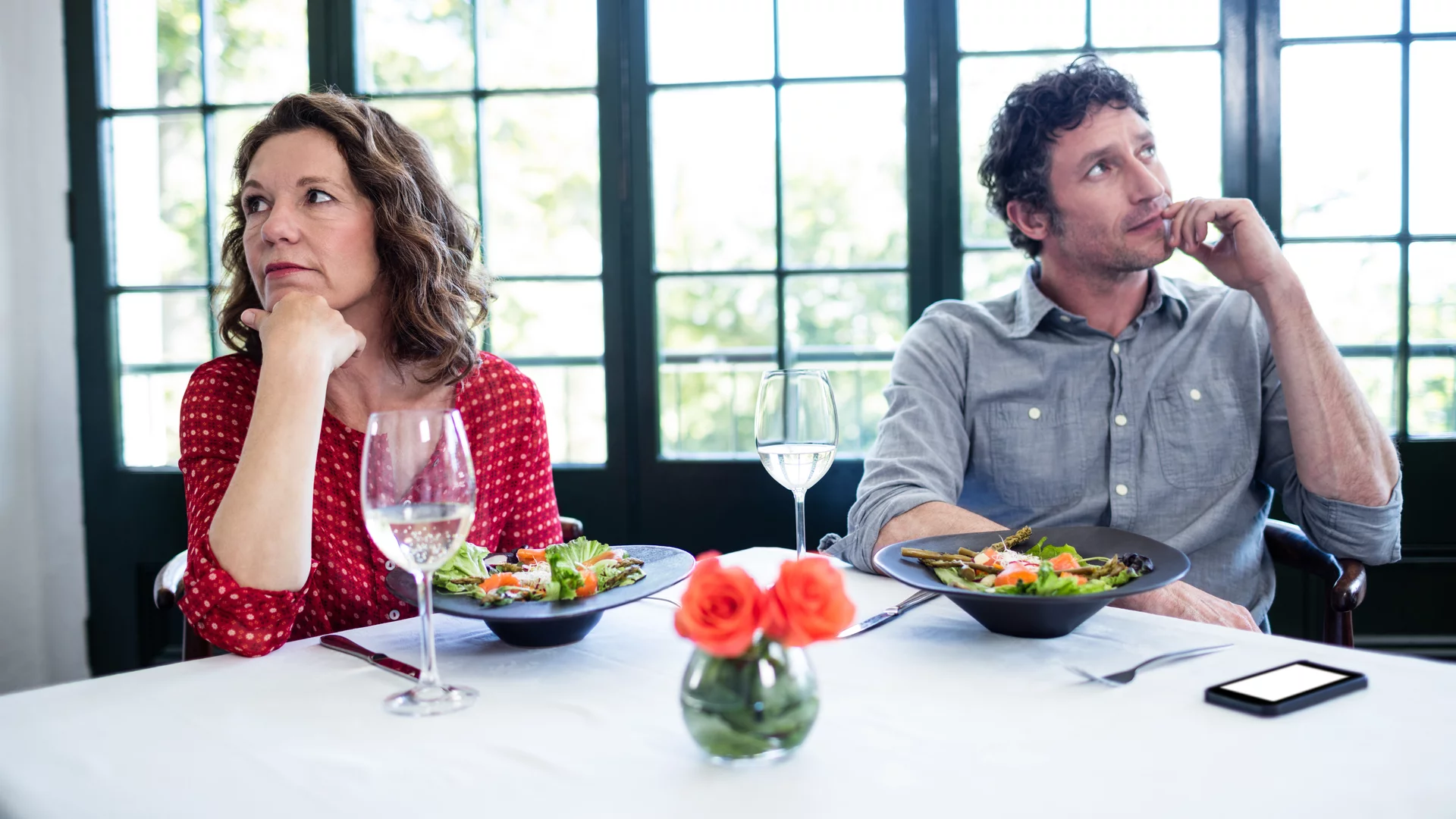 Ein Mann und eine Frau sitzen an einem Tisch mit Salat und Gläsern. Beide wirken nachdenklich und traurig, während sie getrennt voneinander in verschiedene Richtungen schauen. Ein kleines Blumenarrangement steht auf dem Tisch, das eine angenehme Atmosphäre schafft.