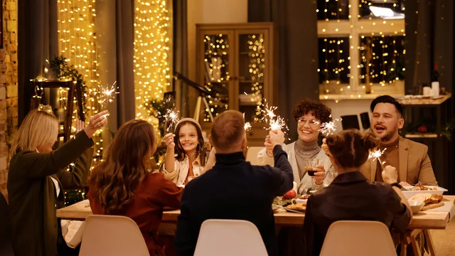 Eine Gruppe von sieben Personen feiert an einem festlich gedeckten Tisch. Jeder hält ein Wunderkerze, die funkelt. Lichterketten schmücken den Hintergrund, und die Atmosphäre ist jubilierend und freundlich. Die Menschen lächeln und scheinen den Moment gemeinsam zu genießen.