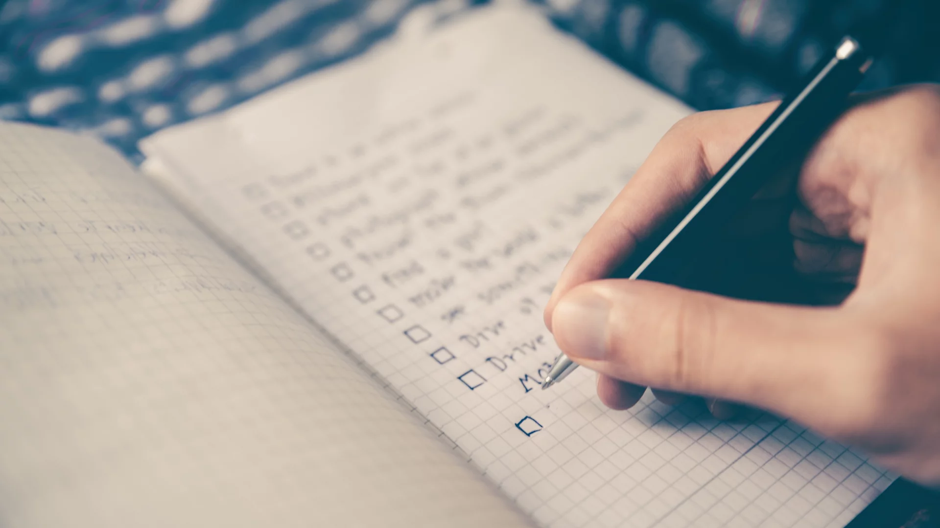 Eine Hand hält einen Stift und kreuzt einen Punkt in einem Notizbuch an, das eine Liste mit Aufgaben oder To-Dos enthält. Die Seite ist mit Kästchen für jede Aufgabe versehen.