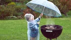 Ein kleines Kind in einem Regenmantel hält einen Regenschirm über einem Grill, während es regnet. Der Grill ist rauchend und auf einem grünen Rasen platziert.
