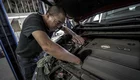 Ein Mann steht neben einem Auto und arbeitet am Motor. Er trägt eine schwarze T-Shirt und eine Brille, während er mit einer Hand an der Maschine hantiert. Im Hintergrund sind Werkstattutensilien sichtbar.