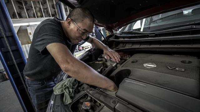 Ein Mann steht neben einem Auto und arbeitet am Motor. Er trägt eine schwarze T-Shirt und eine Brille, während er mit einer Hand an der Maschine hantiert. Im Hintergrund sind Werkstattutensilien sichtbar.