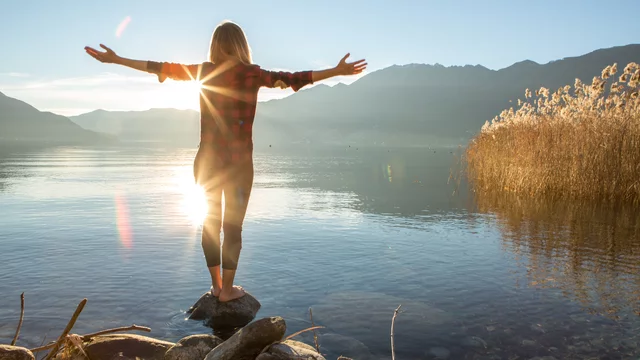 Eine Person steht auf einem Stein am Ufer eines Sees, mit ausgebreiteten Armen. Die Sonne scheint direkt hinter ihr und spiegelt sich im Wasser. Im Hintergrund sind sanfte Hügel und Schilf zu sehen, was eine friedliche, naturverbundene Atmosphäre schafft.