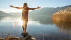 Eine Person steht auf einem Stein am Ufer eines Sees, mit ausgebreiteten Armen. Die Sonne scheint direkt hinter ihr und spiegelt sich im Wasser. Im Hintergrund sind sanfte Hügel und Schilf zu sehen, was eine friedliche, naturverbundene Atmosphäre schafft.