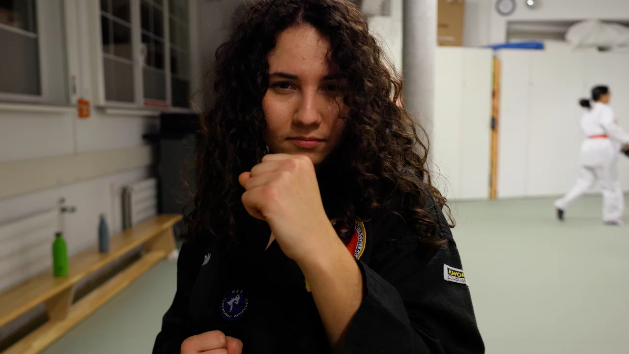 Eine junge Frau mit lockigem Haar trägt einen Karogiganzanzug und zeigt eine Faust, bereit für den Kampfsport. Im Hintergrund sind weitere Personen in weißer Sportkleidung zu sehen, die ebenfalls trainieren. Der Raum wirkt gut beleuchtet und sportlich ausgestattet.