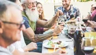 Eine fröhliche Gruppe von Menschen sitzt an einem Tisch, genießt Essen und Trinken. Ein älterer Mann mit Bart hebt sein Glas, während andere lachen und sich unterhalten. Der Tisch ist mit verschiedenen Speisen und Getränken gedeckt, was eine einladende, gesellige Stimmung vermittelt.