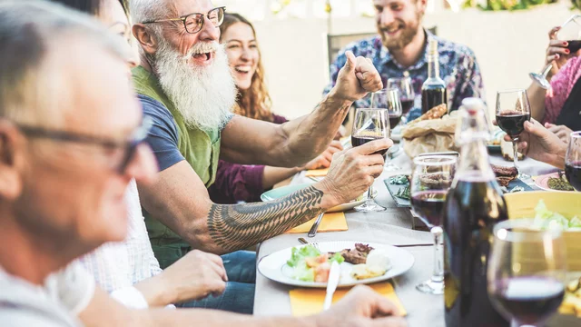 Eine fröhliche Gruppe von Menschen sitzt an einem Tisch, genießt Essen und Trinken. Ein älterer Mann mit Bart hebt sein Glas, während andere lachen und sich unterhalten. Der Tisch ist mit verschiedenen Speisen und Getränken gedeckt, was eine einladende, gesellige Stimmung vermittelt.