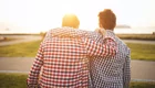 Zwei Männer stehen nebeneinander, mit dem Rücken zur Kamera, und schauen auf das Sonnenlicht am Horizont. Sie tragen karierte Hemden und einer von ihnen hat seinen Arm um den anderen gelegt, was eine freundliche und vertraute Atmosphäre schafft.