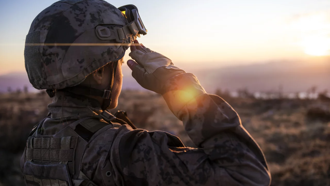 Ein Soldat in militärischer Uniform salutiert mit der rechten Hand. Im Hintergrund steigt die Sonne und taucht die Landschaft in warmes Licht, während der Soldat nachdenklich in die Ferne blickt.