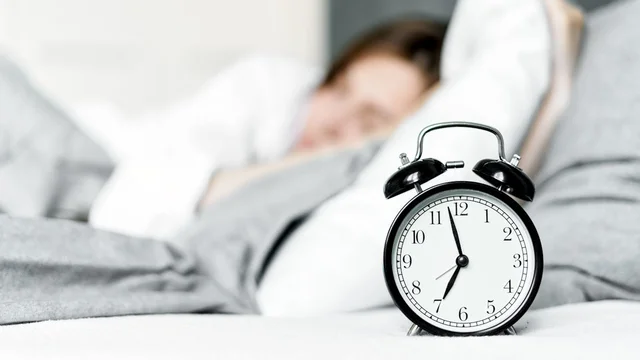 Ein Wecker mit schwarzem Gehäuse und weißen Ziffern zeigt die Uhrzeit an. Im Hintergrund liegt eine Person im Bett, teilweise unscharf. Die Szene vermittelt das Gefühl von Schlaf und Morgenroutine.