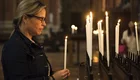 Eine Frau steht an einem Kerzenständer und hält eine brennende Kerze in der Hand. Sie schaut nachdenklich auf die anderen entzündeten Kerzen, die im Hintergrund stehen. Der Ort wirkt feierlich und spirituell.