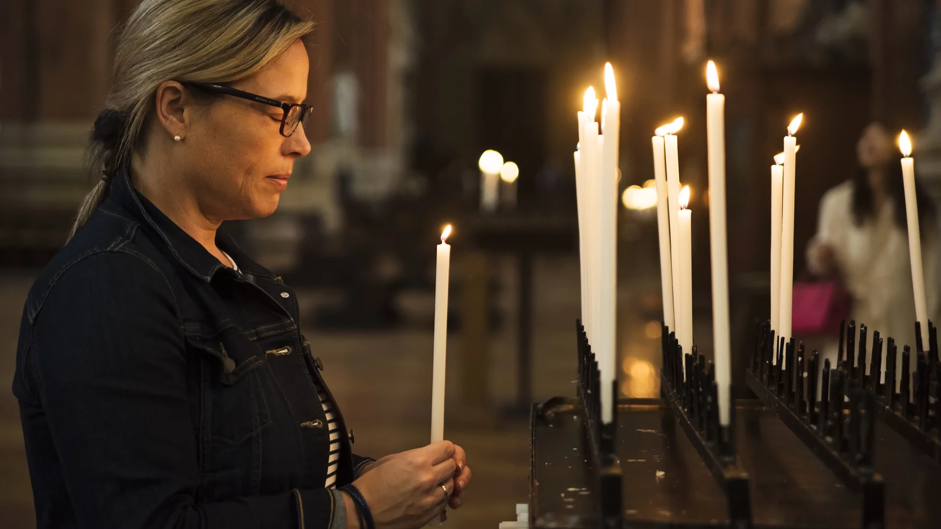Eine Frau steht an einem Kerzenständer und hält eine brennende Kerze in der Hand. Sie schaut nachdenklich auf die anderen entzündeten Kerzen, die im Hintergrund stehen. Der Ort wirkt feierlich und spirituell.