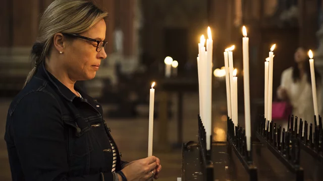 Eine Frau steht an einem Kerzenständer und hält eine brennende Kerze in der Hand. Sie schaut nachdenklich auf die anderen entzündeten Kerzen, die im Hintergrund stehen. Der Ort wirkt feierlich und spirituell.