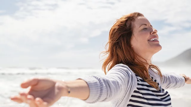 Eine lächelnde Frau mit offenen Armen steht am Strand, während sie den Wind in ihrem Haar spürt. Die Wellen des Meeres sind im Hintergrund zu sehen, und der Himmel ist teilweise bewölkt. Der Ausdruck der Frau zeigt Freude und Freiheit.