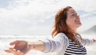 Eine lächelnde Frau mit offenen Armen steht am Strand, während sie den Wind in ihrem Haar spürt. Die Wellen des Meeres sind im Hintergrund zu sehen, und der Himmel ist teilweise bewölkt. Der Ausdruck der Frau zeigt Freude und Freiheit.
