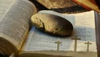 Ein großer Stein liegt auf einer offenen Bibel. Im Hintergrund sind schemenhaft drei Kreuze zu sehen. Die Szene vermittelt ein Gefühl von Spiritualität und Reflexion.