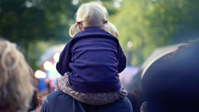 Ein kleines Kind mit blonden Zöpfen sitzt auf den Schultern eines Erwachsenen. Es trägt einen dunklen Pullover und schaut in eine Menschenmenge, umgeben von unscharfen Lichtern und Bäumen im Hintergrund.