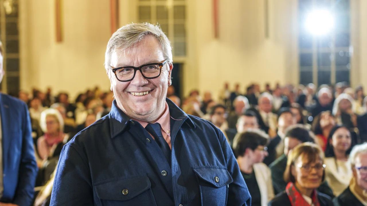 Ein lächelnder Mann mit Brille steht in einem Saal voller Menschen. Im Hintergrund sind zahlreiche Zuschauer zu sehen, die aufmerksam auf die Bühne schauen. Die Beleuchtung ist hell und sorgt für eine angenehme Atmosphäre.