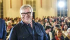 Ein lächelnder Mann mit Brille steht in einem Saal voller Menschen. Im Hintergrund sind zahlreiche Zuschauer zu sehen, die aufmerksam auf die Bühne schauen. Die Beleuchtung ist hell und sorgt für eine angenehme Atmosphäre.