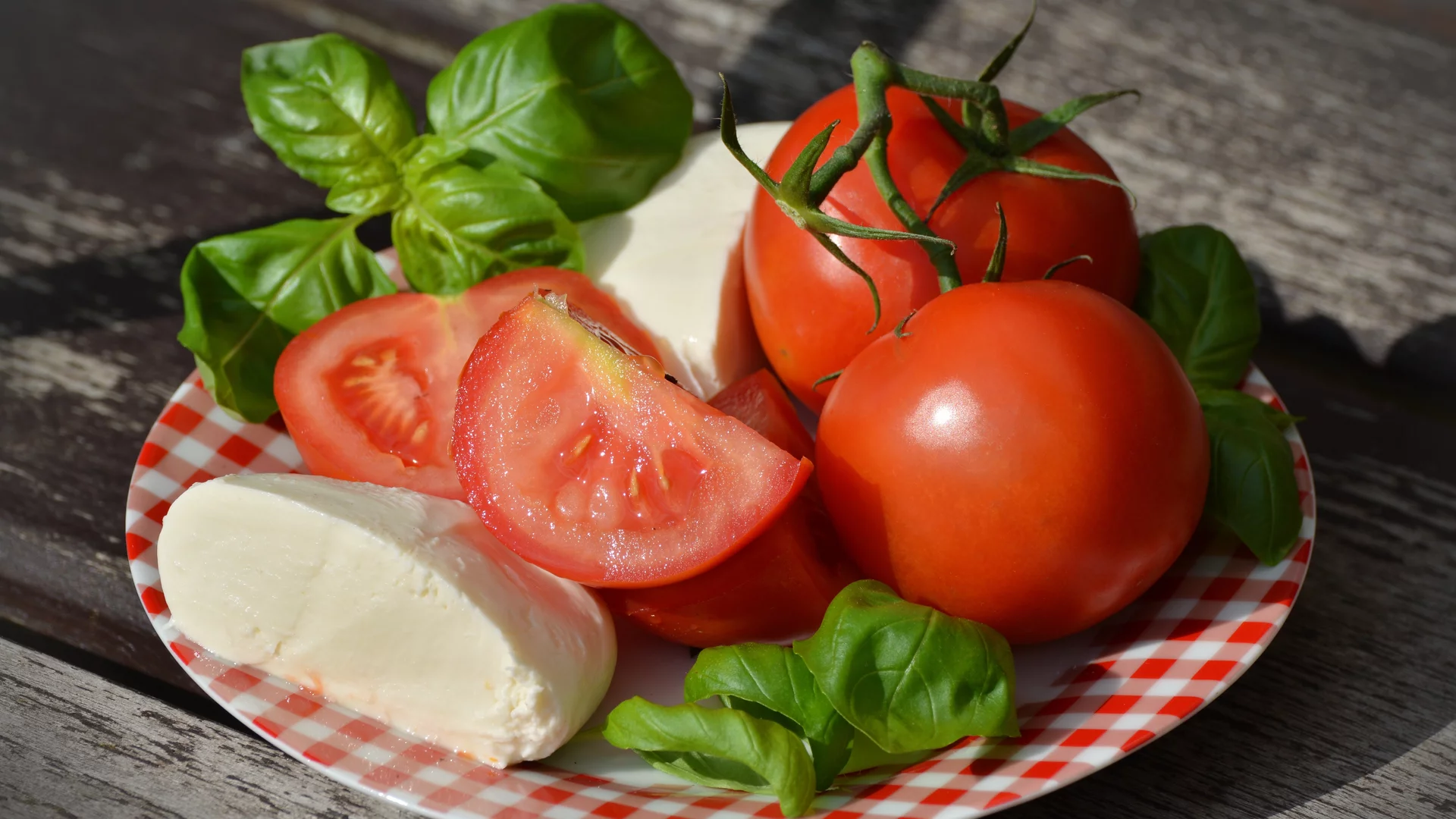 Eine flache, karierte Platte mit frischen, roten Tomaten, halbierten Tomaten, Mozzarella und frischem Basilikum. Die Zutaten sind farbenfrohe und appetitliche Lebensmittel, die für einen Salat oder eine Vorspeise geeignet sind.