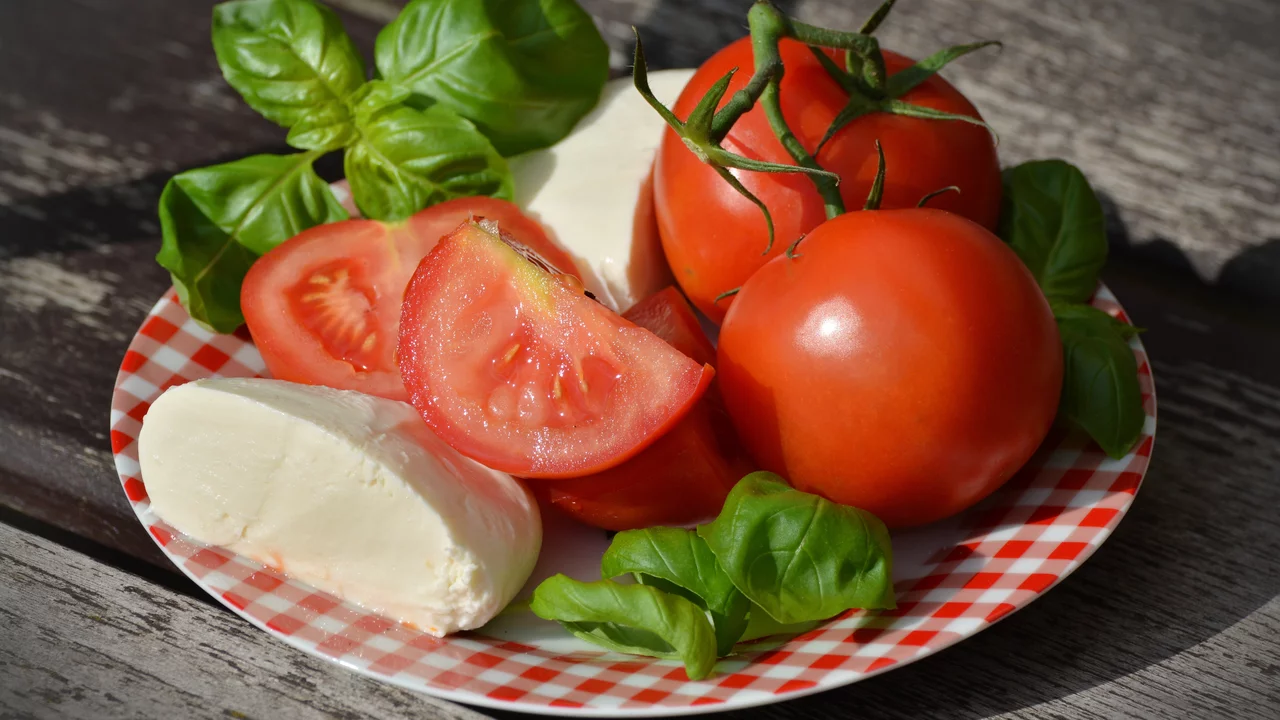 Eine flache, karierte Platte mit frischen, roten Tomaten, halbierten Tomaten, Mozzarella und frischem Basilikum. Die Zutaten sind farbenfrohe und appetitliche Lebensmittel, die für einen Salat oder eine Vorspeise geeignet sind.