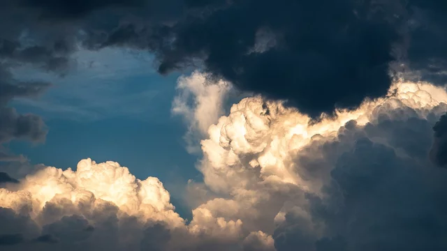 Eine dramatische Wolkenlandschaft ist zu sehen, mit dunklen, bedrohlichen Wolken und leuchtend weißen Wolken, die lichtdurchflutet sind. Der Himmel zeigt verschiedene Blautöne und gibt einen Eindruck von gewitterartigem Wetter.