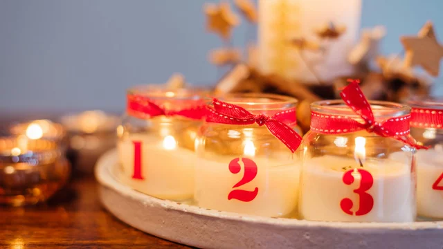 Eine festliche Anordnung von vier Kerzen mit roten Schleifen, nummeriert von 1 bis 4. Sie stehen auf einem runden Tablett, umgeben von einer gemütlichen Atmosphäre. Im Hintergrund brennen weitere Kerzen, die eine warme Beleuchtung erzeugen.