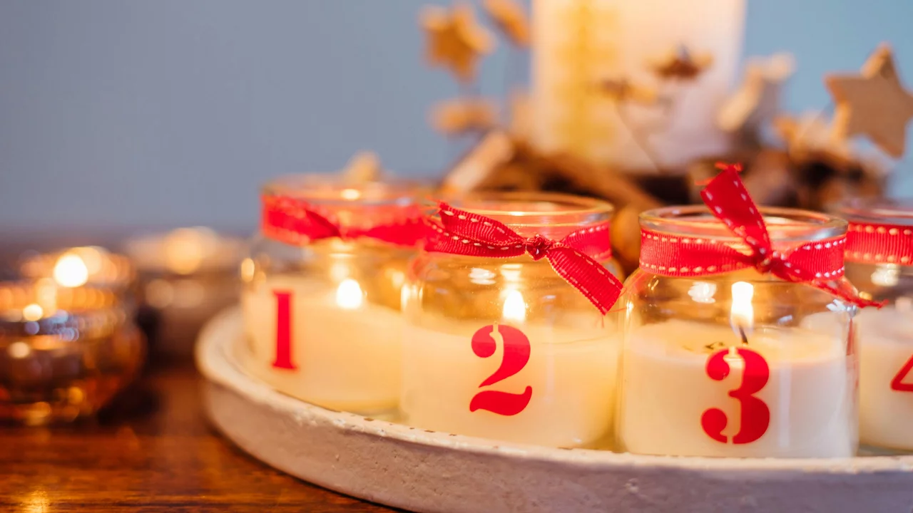 Eine festliche Anordnung von vier Kerzen mit roten Schleifen, nummeriert von 1 bis 4. Sie stehen auf einem runden Tablett, umgeben von einer gemütlichen Atmosphäre. Im Hintergrund brennen weitere Kerzen, die eine warme Beleuchtung erzeugen.