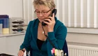 Eine Frau mit blonden, kurzen Haaren sitzt an einem Schreibtisch und telefoniert. Sie trägt einen blauen Pullover und macht Notizen auf einem Blatt Papier. Auf dem Tisch befinden sich Stifte und Bürobedarf.