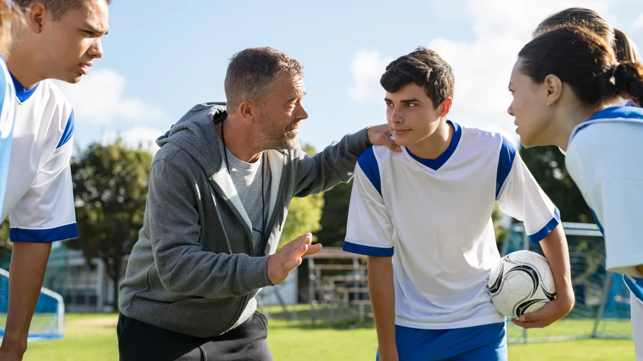 Ein Trainer steht mit einer Gruppe junger Fußballspieler auf einem Spielfeld. Er erklärt etwas eindringlich, während die Spieler aufmerksam zuhören und einen Fußball halten. Der Trainer trägt einen grauen Hoodie, die Spieler sind in weißen und blauen Trikots gekleidet.