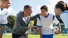 Ein Trainer steht mit einer Gruppe junger Fußballspieler auf einem Spielfeld. Er erklärt etwas eindringlich, während die Spieler aufmerksam zuhören und einen Fußball halten. Der Trainer trägt einen grauen Hoodie, die Spieler sind in weißen und blauen Trikots gekleidet.