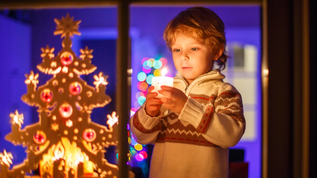 Ein Junge steht vor einem Fenster und hält eine Kerze in der Hand. Hinter ihm ist ein festlich geschmückter Weihnachtsbaum mit bunten Lichtern. Die Szene strahlt eine warme, weihnachtliche Atmosphäre aus.