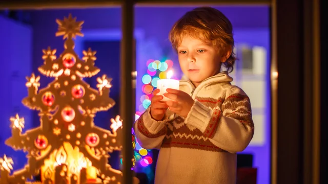 Ein Junge steht vor einem Fenster und hält eine Kerze in der Hand. Hinter ihm ist ein festlich geschmückter Weihnachtsbaum mit bunten Lichtern. Die Szene strahlt eine warme, weihnachtliche Atmosphäre aus.