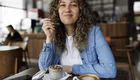 Eine junge Frau mit lockigem Haar sitzt in einem Café und lächelt, während sie einen Löffel mit einem Dessert hält. Auf dem Tisch vor ihr sind ein schmackhaftes Dessert in einer kleinen Schüssel und eine Tasse Kaffee zu sehen.