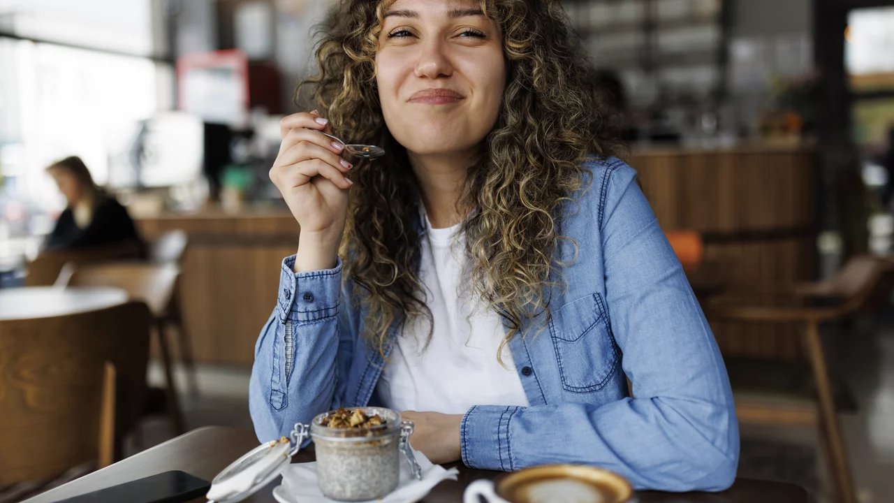 Eine junge Frau mit lockigem Haar sitzt in einem Café und lächelt, während sie einen Löffel mit einem Dessert hält. Auf dem Tisch vor ihr sind ein schmackhaftes Dessert in einer kleinen Schüssel und eine Tasse Kaffee zu sehen.