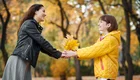 Eine Frau in schwarzer Lederjacke und ein Mädchen in einer gelben Jacke stehen sich gegenüber. Das Mädchen hält einen Strauß aus bunten Herbstblättern. Beide lächeln sich an, umgeben von einer herbstlichen Parklandschaft mit orangefarbenen und gelben Blättern.
