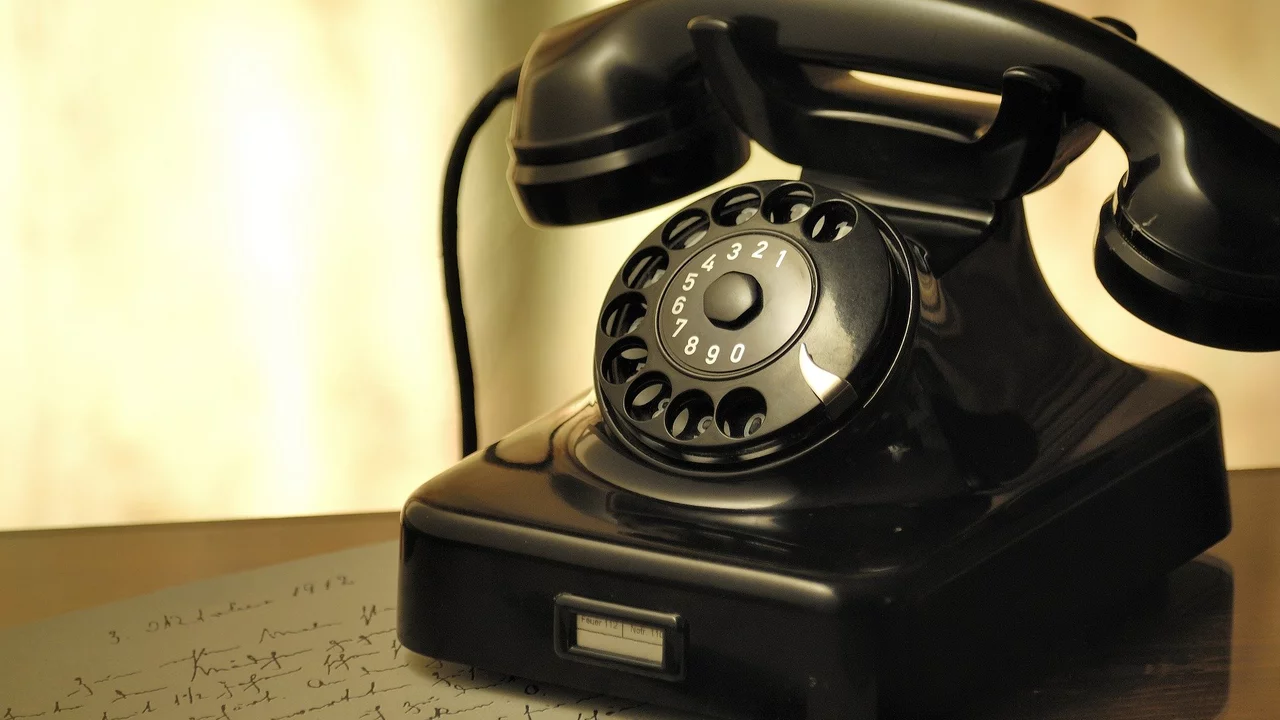 Ein schwarzes, retro Telefon mit Wählscheibe steht auf einem Tisch neben einem Blatt mit handschriftlichen Notizen und einem Füller. Das Bild vermittelt ein Gefühl von Nostalgie und Kommunikation.
