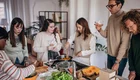 Sechs Personen stehen in einer modernen Küche und bereiten gemeinsam Essen zu. Sie schneiden Gemüse, kochen und haben Getränke in Gläsern. Die Atmosphäre ist freundlich und kollaborativ, und es gibt frische Zutaten auf der Arbeitsfläche.
