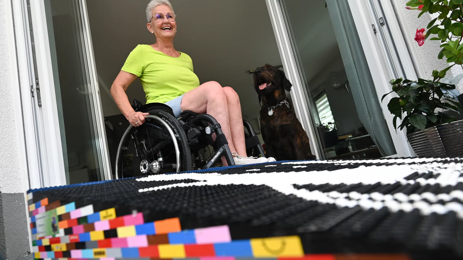 Eine ältere Frau - Rita Ebel - in einem Rollstuhl lächelt, während sie in ihre Wohnung rollt. Neben ihr steht ein Hund. Der Türbereich ist farbenfroh mit einem bunten, spielerischen Lego-Teppich gestaltet, der einen einladenden Eindruck vermittelt.