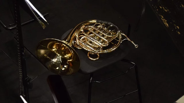 Ein glänzendes Horn liegt auf einem Stuhl in einem dunklen Raum. Der Boden ist schwarz, und im Hintergrund sind einige Notenständer sichtbar. Das Horn scheint bereit zu sein für eine musikalische Darbietung.