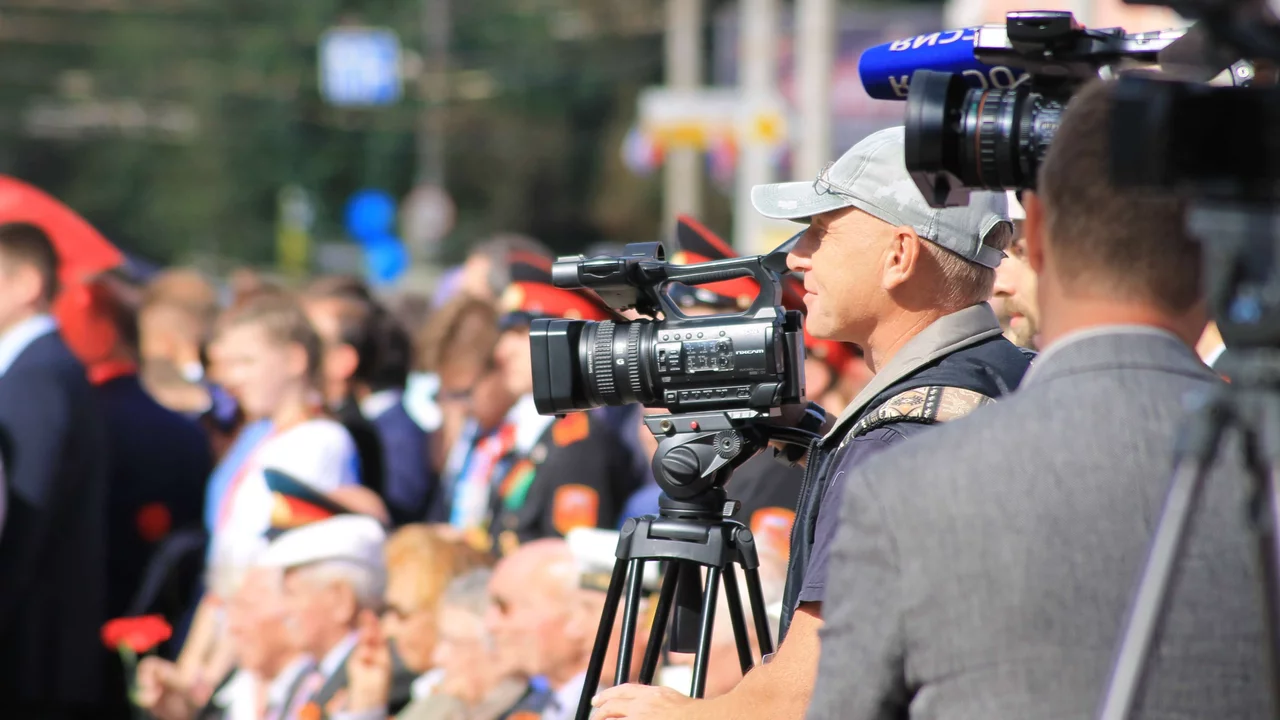 Die Aufnahme zeigt einen Kameramann, der eine Veranstaltung filmt. Im Hintergrund sind Menschen in festlicher Kleidung zu sehen, wahrscheinlich bei einer Feier oder Zeremonie. Die Atmosphäre wirkt lebhaft und die Umgebung ist hell beleuchtet.