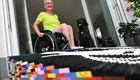Eine ältere Frau sitzt in einem Rollstuhl und lächelt, während sie auf einer bunten LEGO-Matte vor einer offenen Tür steht. Ein Hund ist im Hintergrund zu sehen. Die Szene vermittelt Freude und Offenheit.