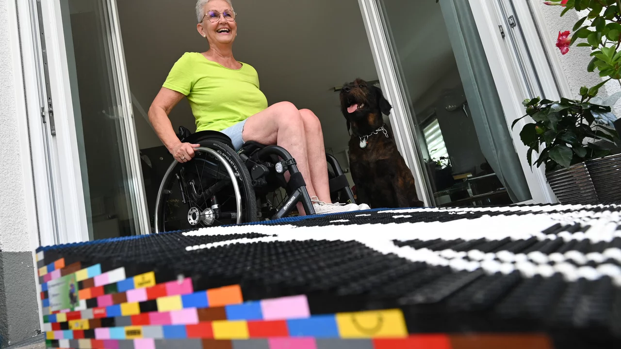 Eine ältere Frau sitzt in einem Rollstuhl und lächelt, während sie auf einer bunten LEGO-Matte vor einer offenen Tür steht. Ein Hund ist im Hintergrund zu sehen. Die Szene vermittelt Freude und Offenheit.