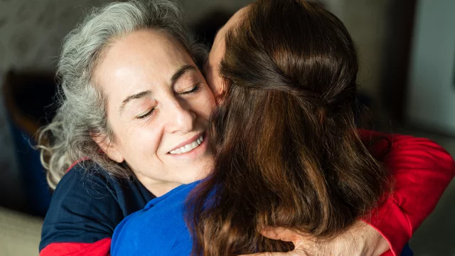 Zwei Frauen umarmen sich herzlich, lächelnd und zufrieden. Die eine hat graue Haare und trägt ein dunkelblaues Oberteil, während die andere lange braune Haare hat und ein blaues Shirt trägt. Der Moment drückt Nähe und Freude aus.