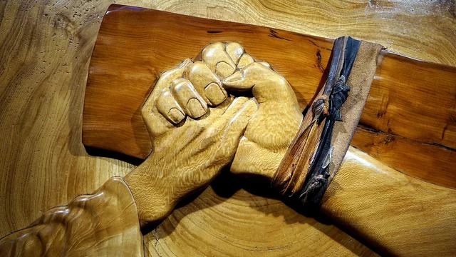 Das Bild zeigt eine kunstvoll geschnitzte Darstellung von zwei Händen, die festen Halt ineinander halten. Die Hände sind aus Holz und vermitteln ein Gefühl von Zusammenhalt und Verbundenheit. Ein Lederband umschlingt das Handgelenk einer der Hände.