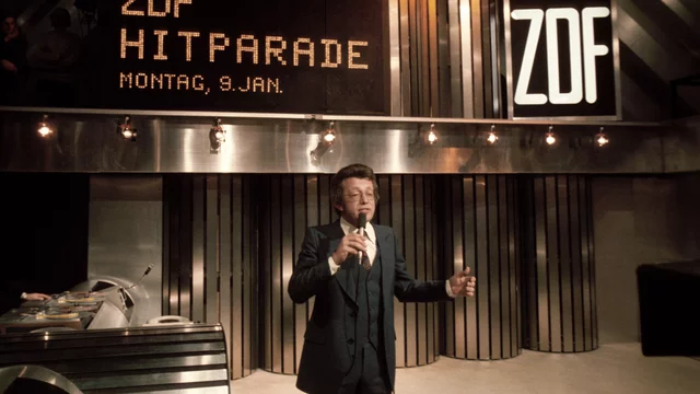 Ein Moderator steht auf einer Bühne, steht im Anzug und spricht ins Mikrofon. Im Hintergrund sind die Worte "ZDF HITPARADE" und das Datum "Montag, 9. Jan." auf einer Leinwand zu sehen. Die Kulisse ist modern und stimmungsvoll beleuchtet.