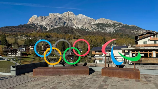 Zwei Skulpturen vor einer Berglandschaft zeigen die olympischen Ringe in verschiedenen Farben und das Paralympics-Logo. Der klare Himmel und die umgebenden Berge schaffen eine beeindruckende Kulisse.